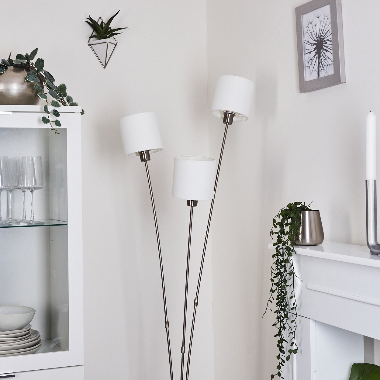 Ein Raum mit einem weißen Schrank, einer dreiflammigen Stehlampe und einem Bilderrahmen. Pflanzen und Geschirr sind ebenfalls zu sehen.
