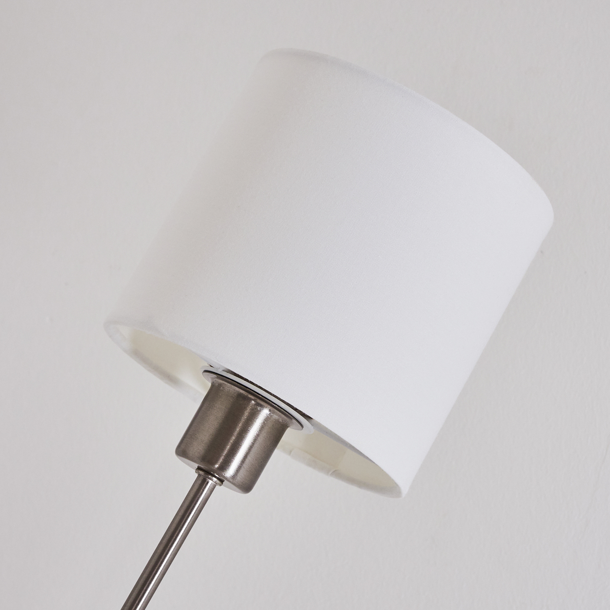 Ein weißer Lampenschirm ist gegen eine weiße Wand geneigt. Eine silberne Metallstange stützt den Lampenschirm.