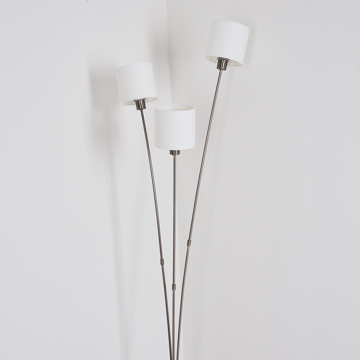 Eine Stehlampe mit drei weißen Lampenschirmen und Metallstielen steht an einer weißen Wand. Die Lampe steht in der Ecke des Raumes.