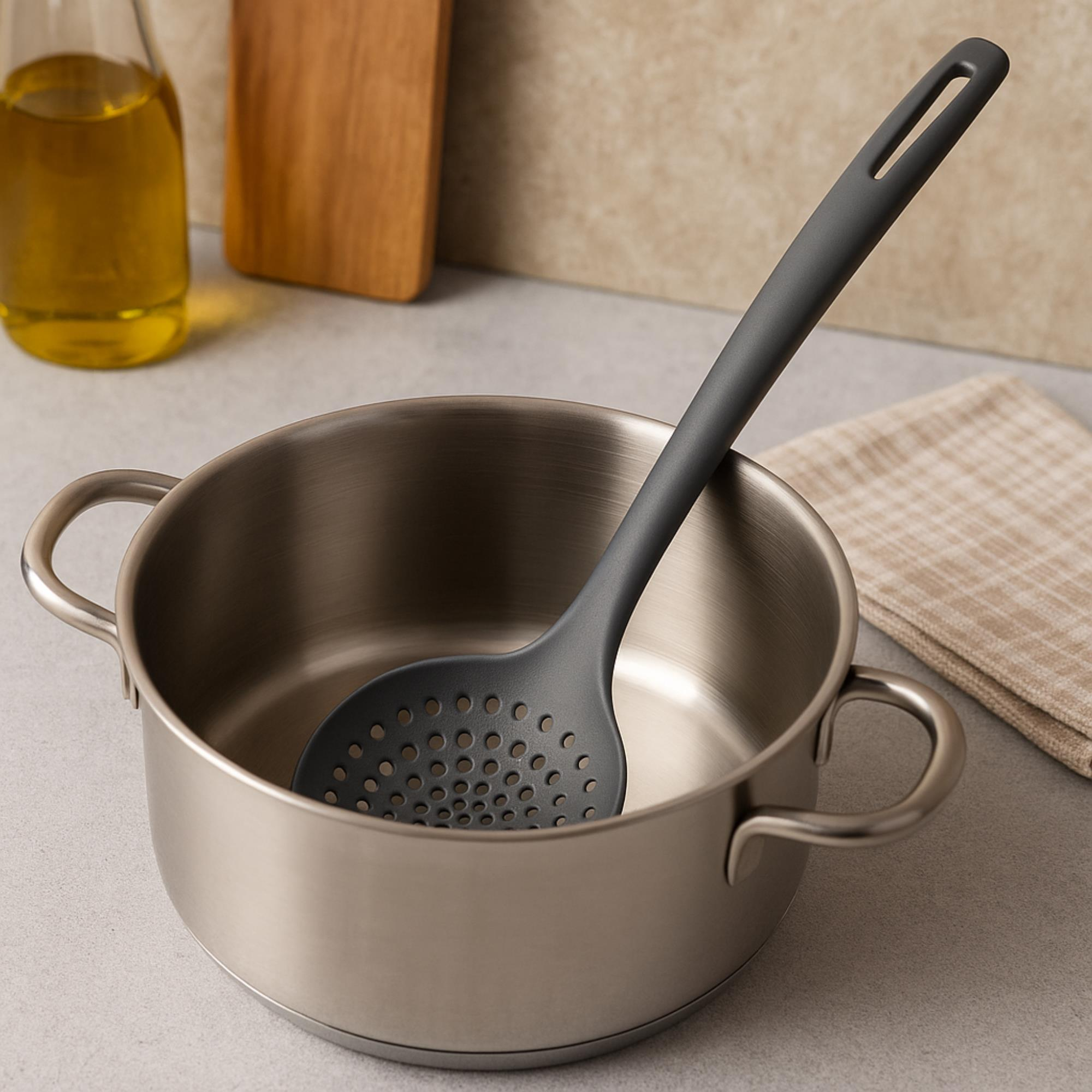 Una cuchara ranurada gris está dentro de una olla de acero inoxidable. Una botella de aceite y una tabla de cortar están en el fondo.