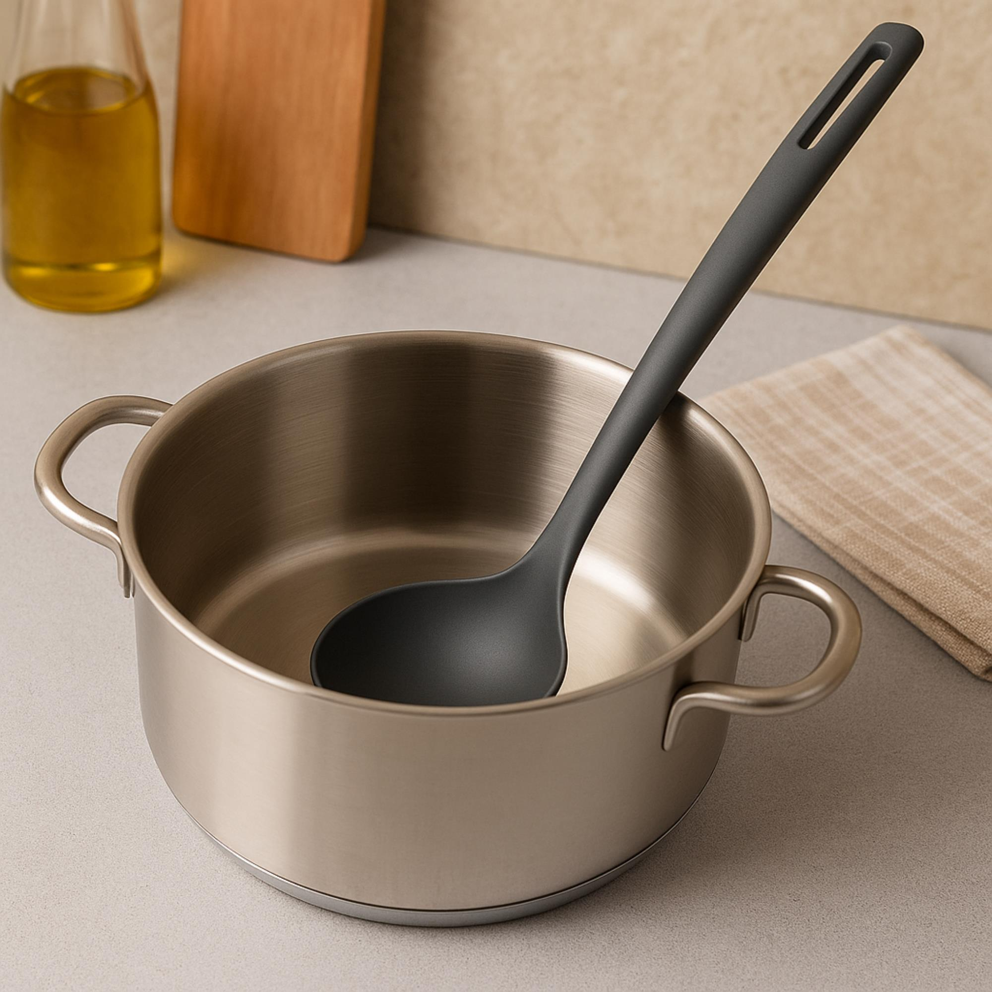 Una olla de acero inoxidable con un cucharón gris oscuro dentro. Aceite y una tabla de cortar en el fondo.