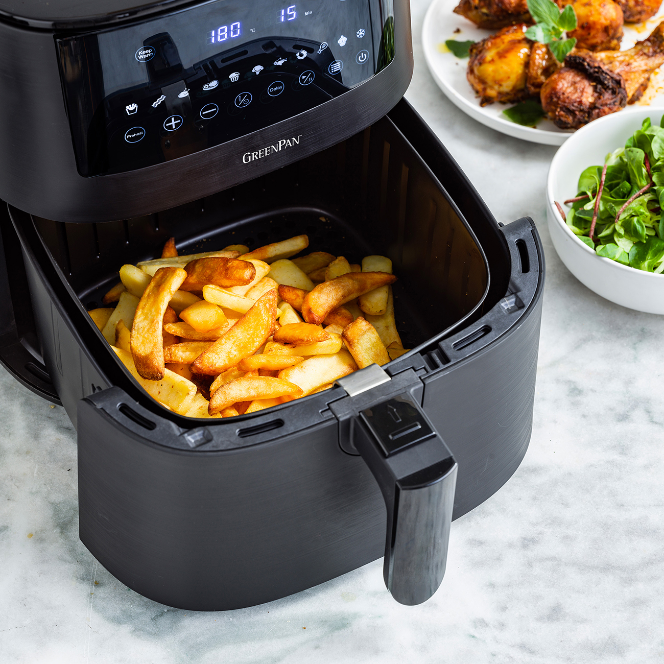 Friggitrice ad aria nera con patatine fritte cotte. Pollo e insalata su piatti sullo sfondo. Il display digitale mostra le impostazioni.