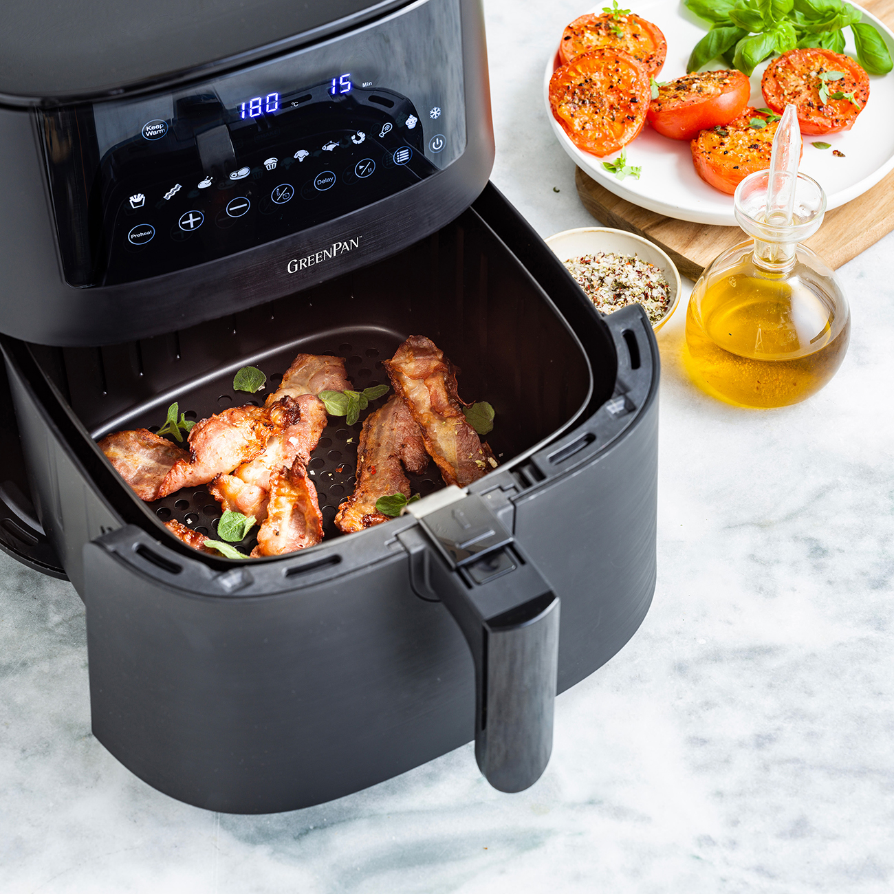 Una friggitrice ad aria con pancetta. Pomodori e olio nelle vicinanze. La friggitrice è nera, con display digitale.