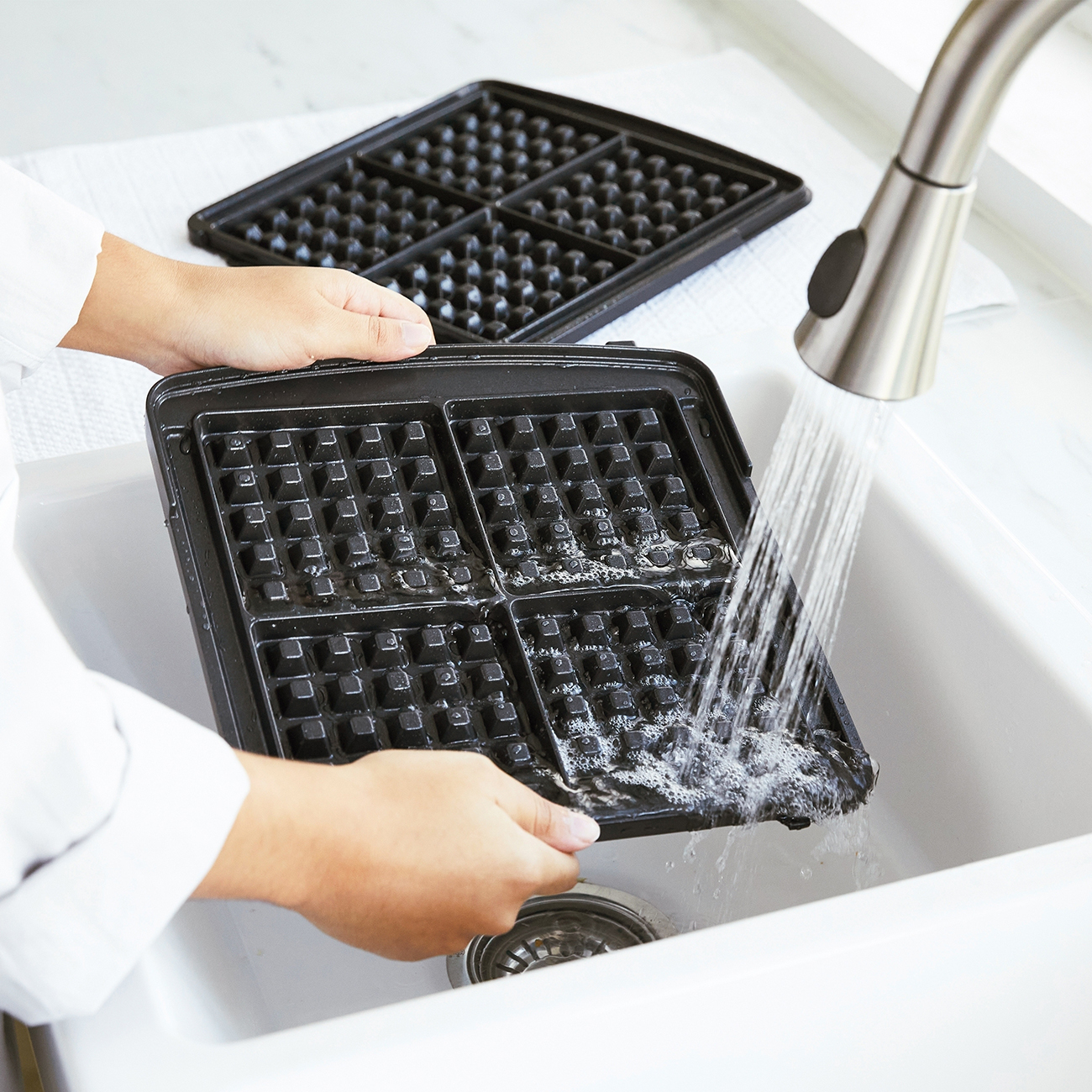 Eine Person wäscht ein schwarzes Waffeleisen unter einem Wasserhahn in einem weißen Waschbecken. Ein weiteres Waffeleisenteil liegt daneben.