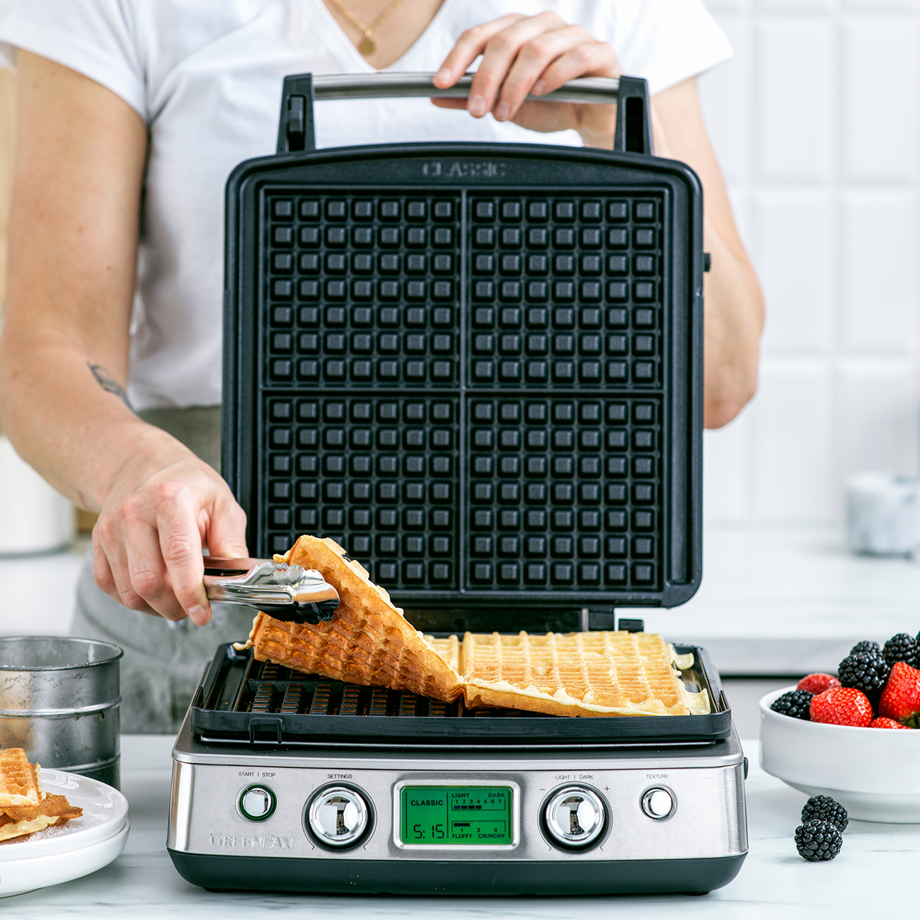 Eine Person entnimmt mit einer Zange einen Waffel aus dem Waffeleisen. Beeren sind in einer weißen Schüssel rechts.