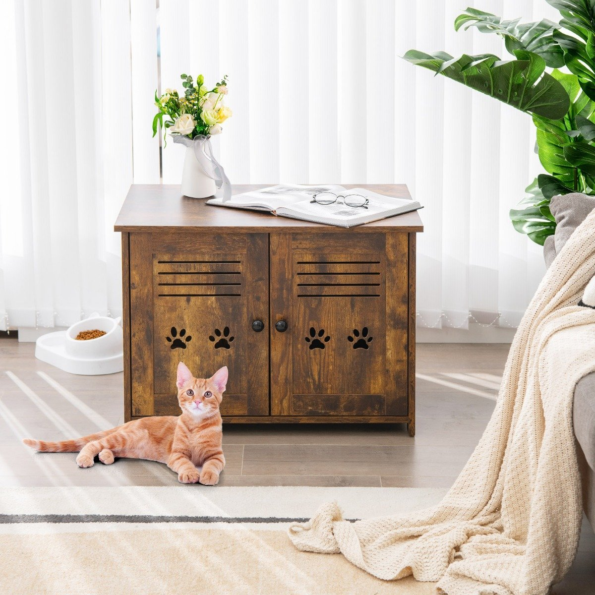 Un gato atigrado naranja yace cerca de un mueble de madera. El mueble tiene huellas de patas. Un jarrón de flores blanco y un libro están encima.
