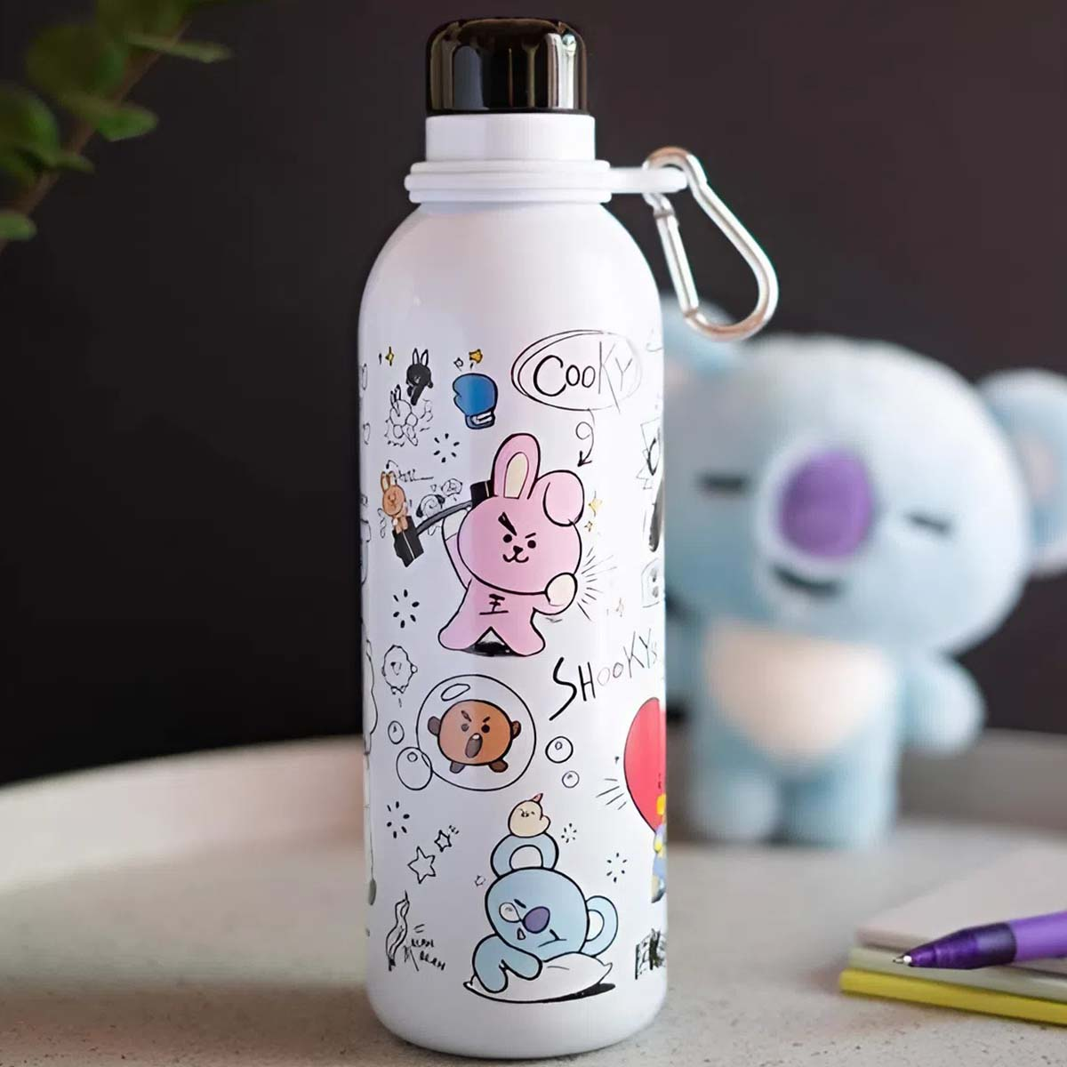 Weiße Wasserflasche mit Cartoonfiguren. Koala-Plüschtier im Hintergrund. Stift, Notizbuch und Pflanze auf dem Tisch.