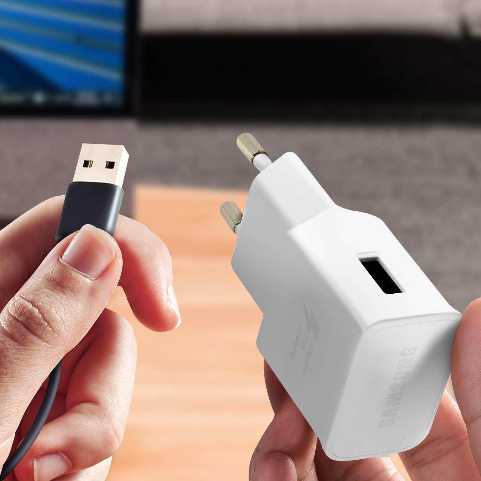 Une main tient un câble USB et l'autre un chargeur Samsung blanc. Arrière-plan : ordinateur flou.