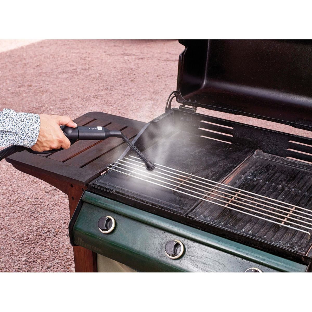 Osoba czyści czarnego grilla parą. Grill stoi na drewnianym stole. Tło to ziemia pokryta kamykami.