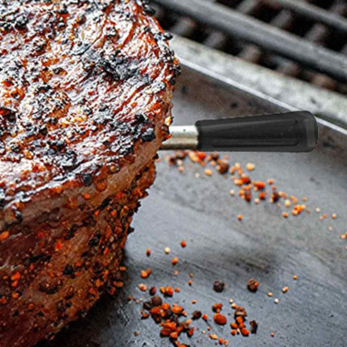 Nahaufnahme eines gegrillten Steaks mit einem Temperaturfühler. Das Steak ist mit Gewürzen gewürzt, auf einer Metalloberfläche.