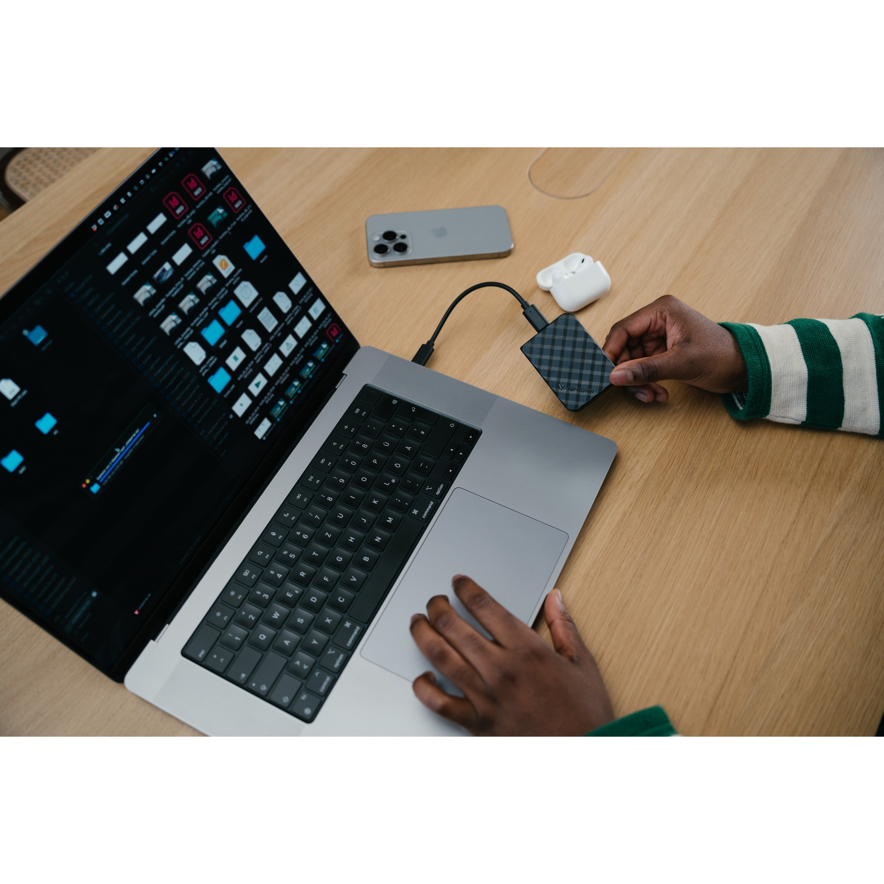 Una persona tiene in mano un disco rigido esterno nero collegato a un laptop. Un iPhone e auricolari sono vicini.