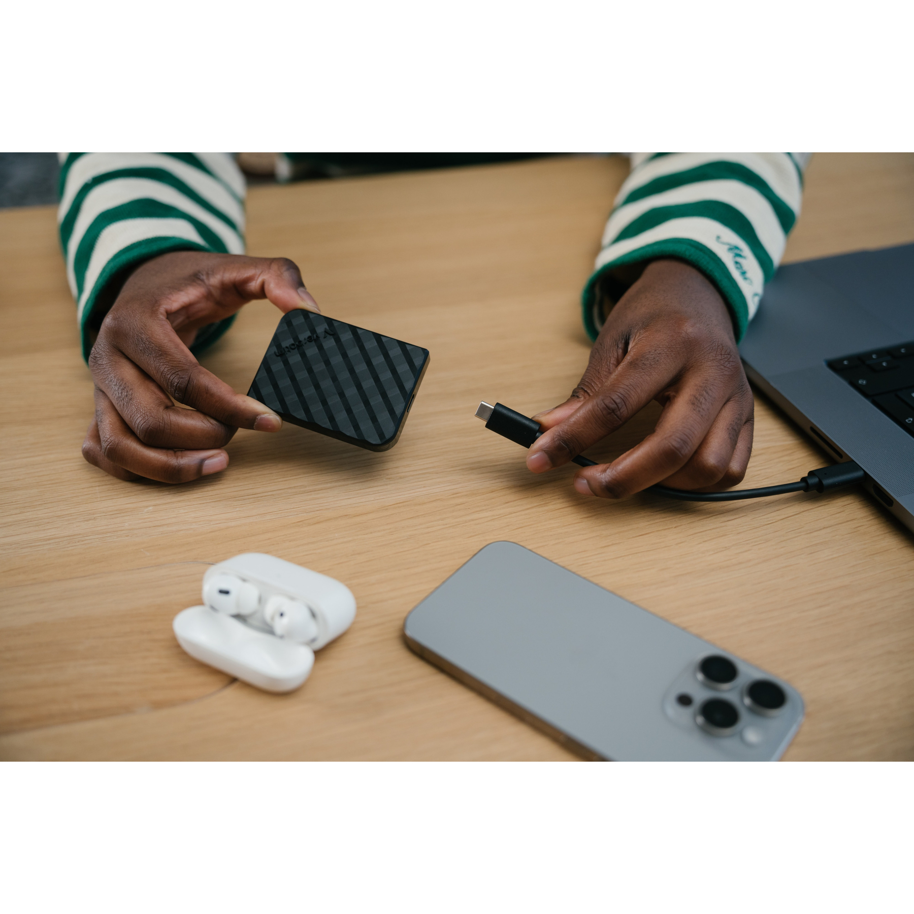 Mani che tengono un disco rigido esterno nero e un cavo USB-C, con un laptop, telefono e auricolari su un tavolo di legno.