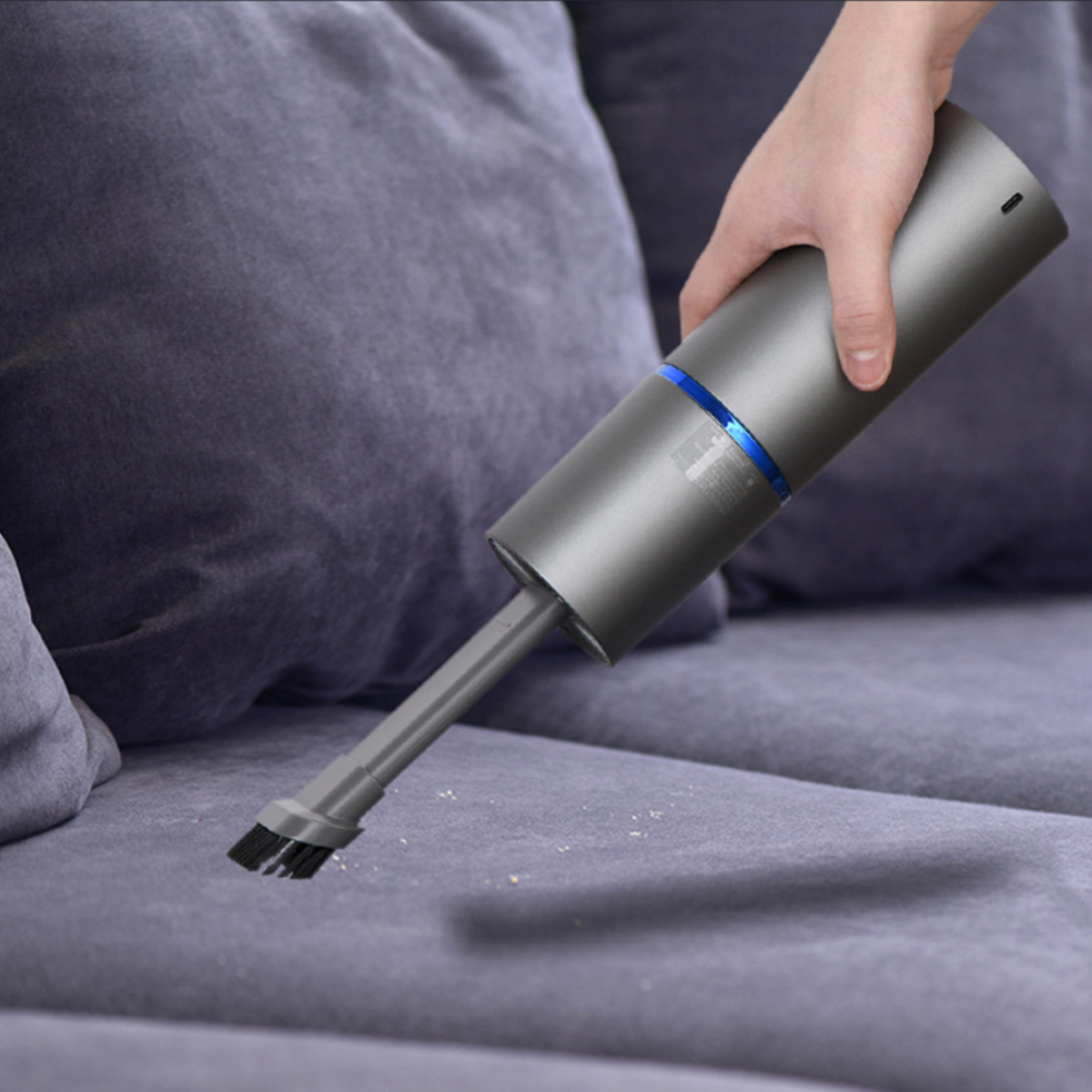 Eine Hand hält einen grauen Handstaubsauger über einem grauen Sofa und reinigt es. Der Staubsauger hat einen blauen Ring und einen Bürstenaufsatz.