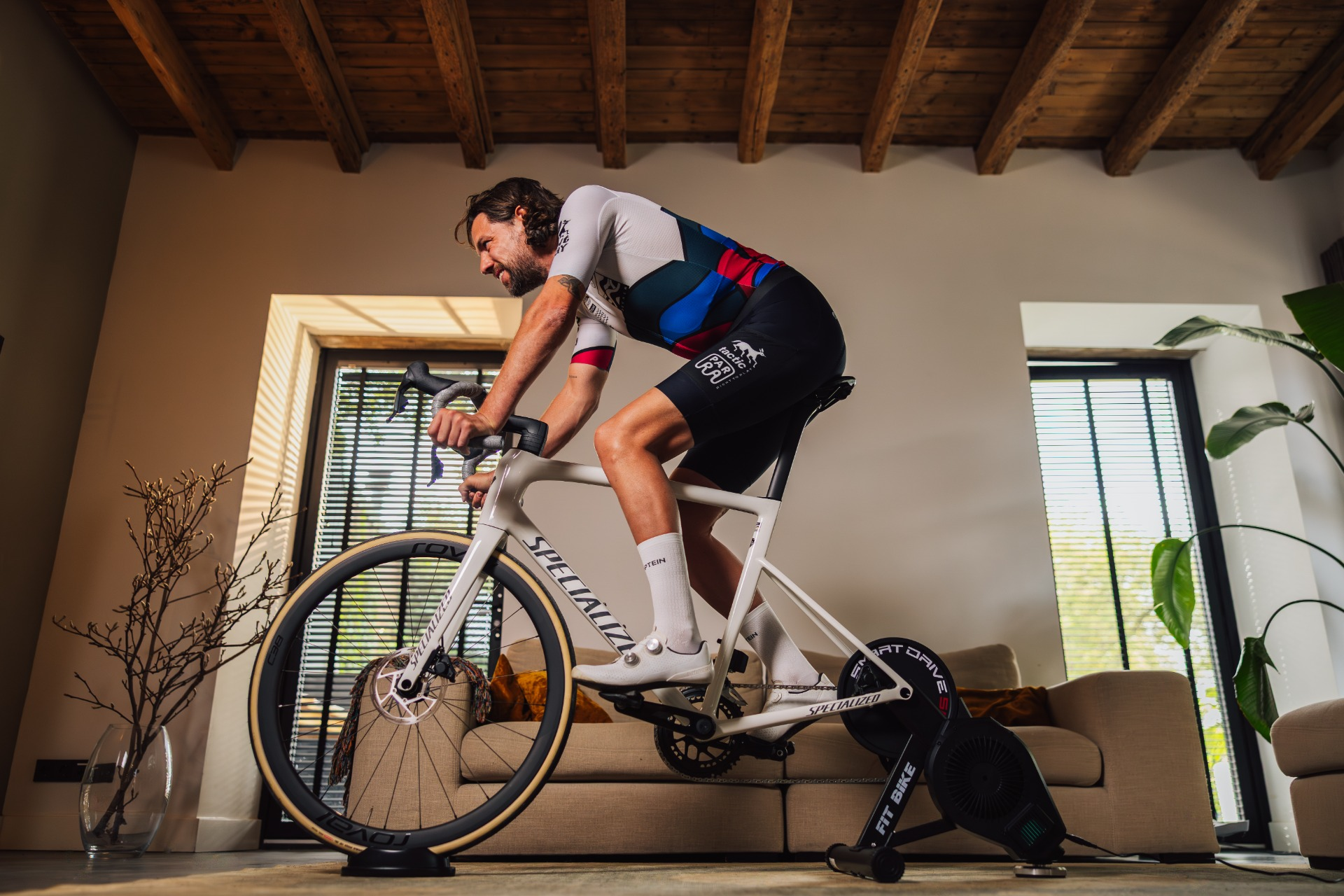 Man op een witte fiets binnenshuis. Hij draagt een wielershirt, een zwarte korte broek en witte sokken. De kamer heeft een houten plafond, een bank en ramen.