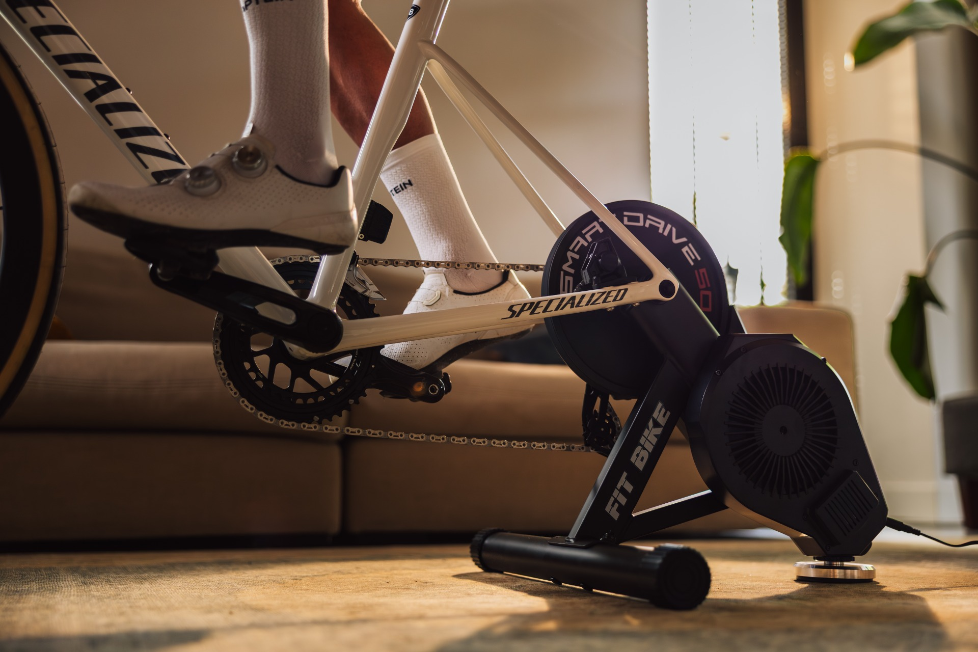 Een persoon fietst binnen. Witte fiets met witte schoenen. De fiets is aangesloten op een zwarte Fit Bike trainer. Zonnestralen op de achtergrond.