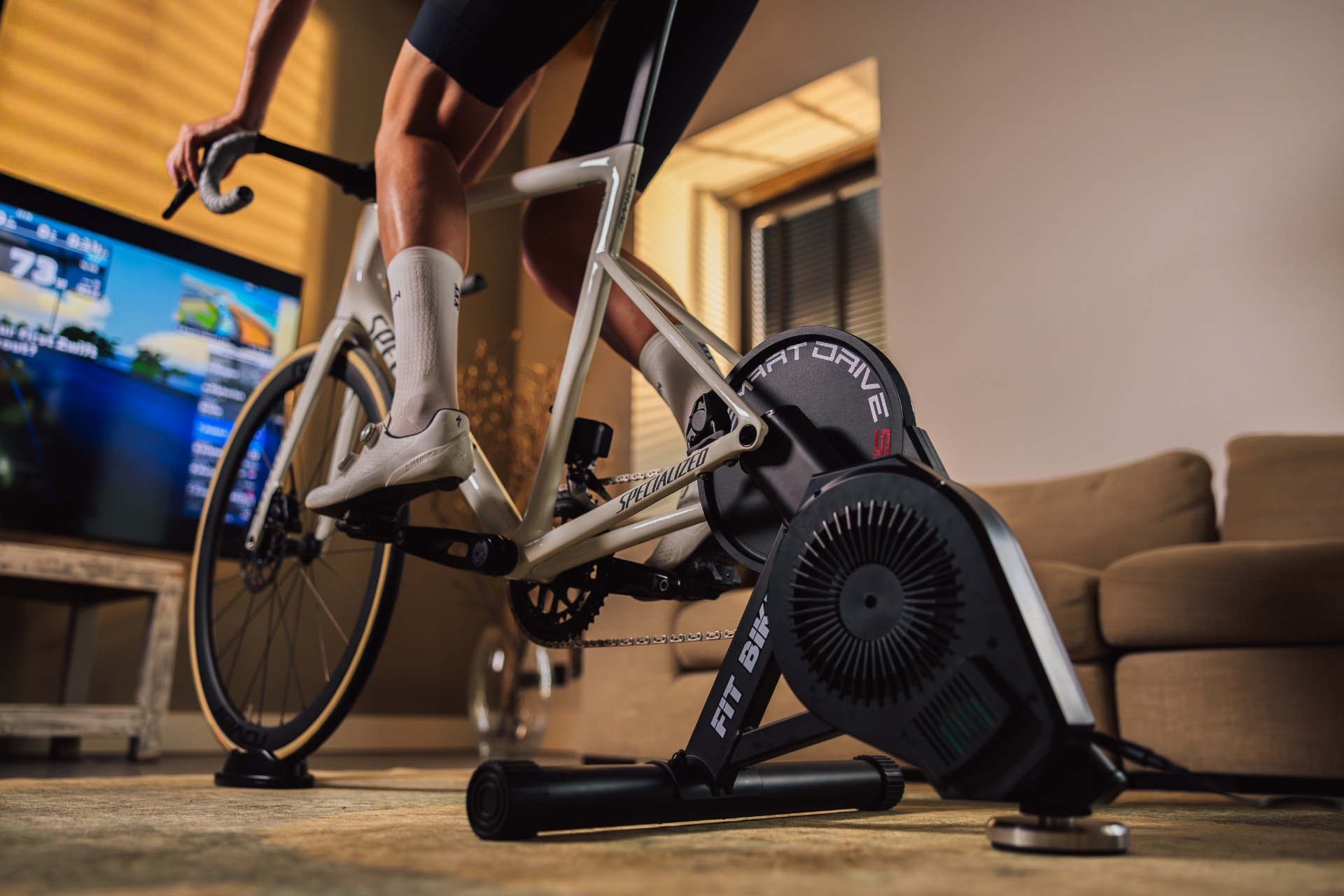 Fietser op witte fiets gebruikt Smart Drive, binnen. Een tv-scherm toont gegevens. De achtergrond toont een bank en een raam.