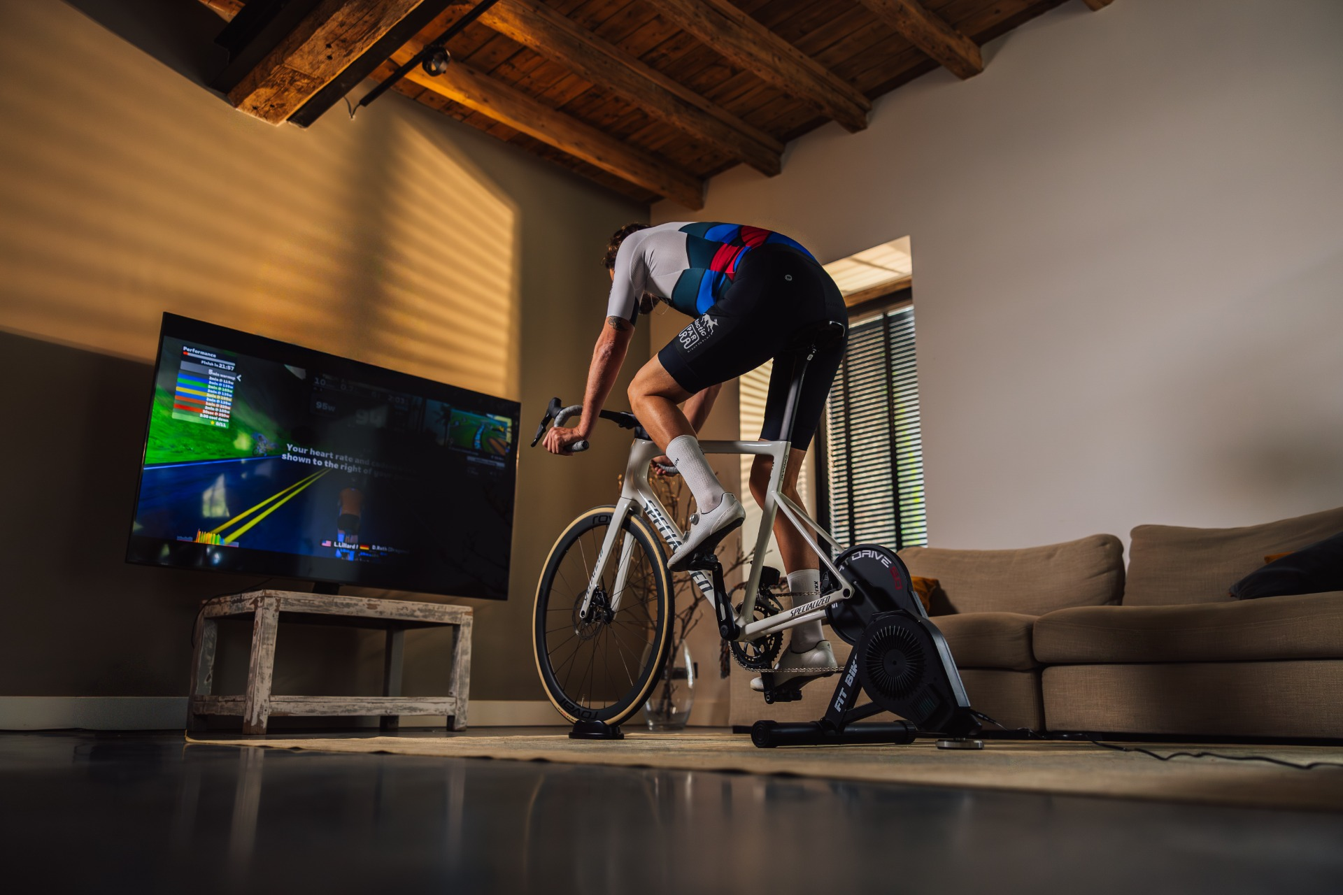 Een fietser in een home gym. Hij zit op een witte fiets die is aangesloten op een trainer en kijkt naar een scherm. Woonkamer met een bank zichtbaar.
