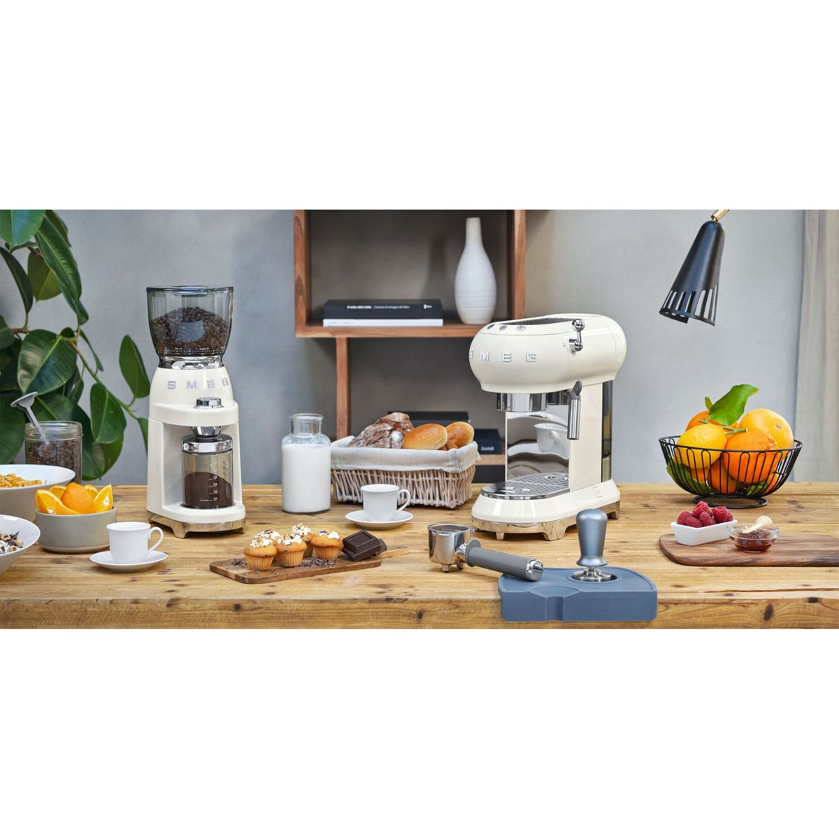 Eine Küchenszene mit Kaffeemaschine, Mühle, Früchten, Gebäck und Brot auf einem Holztisch. Im Hintergrund eine Pflanze.