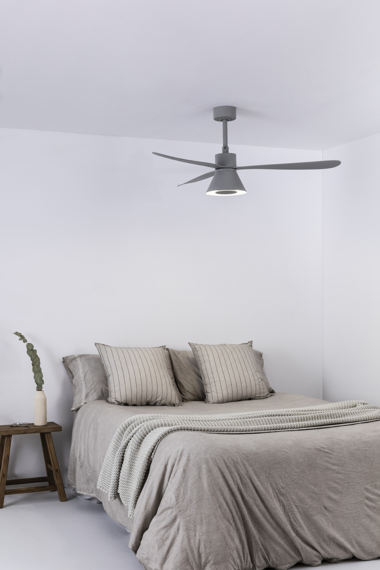 Ein Schlafzimmer mit Bett, zwei Kissen, einer Decke und einem grauen Deckenventilator. Ein kleiner Holztisch steht neben dem Bett.