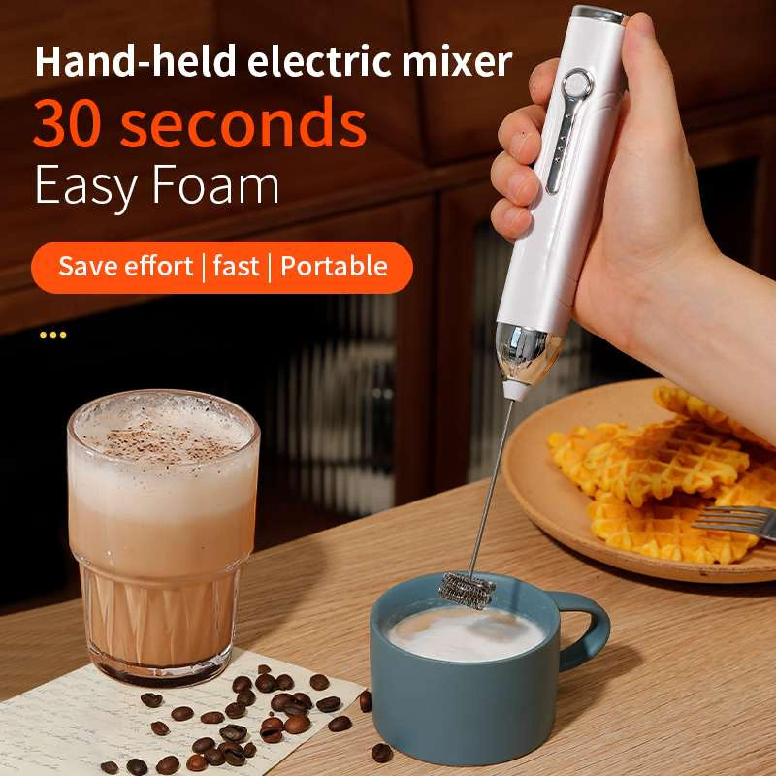 Eine Hand, die einen elektrischen Mixer über einer blauen Schaumtasse hält. Ein Glas Kaffee und Waffeln stehen in der Nähe.