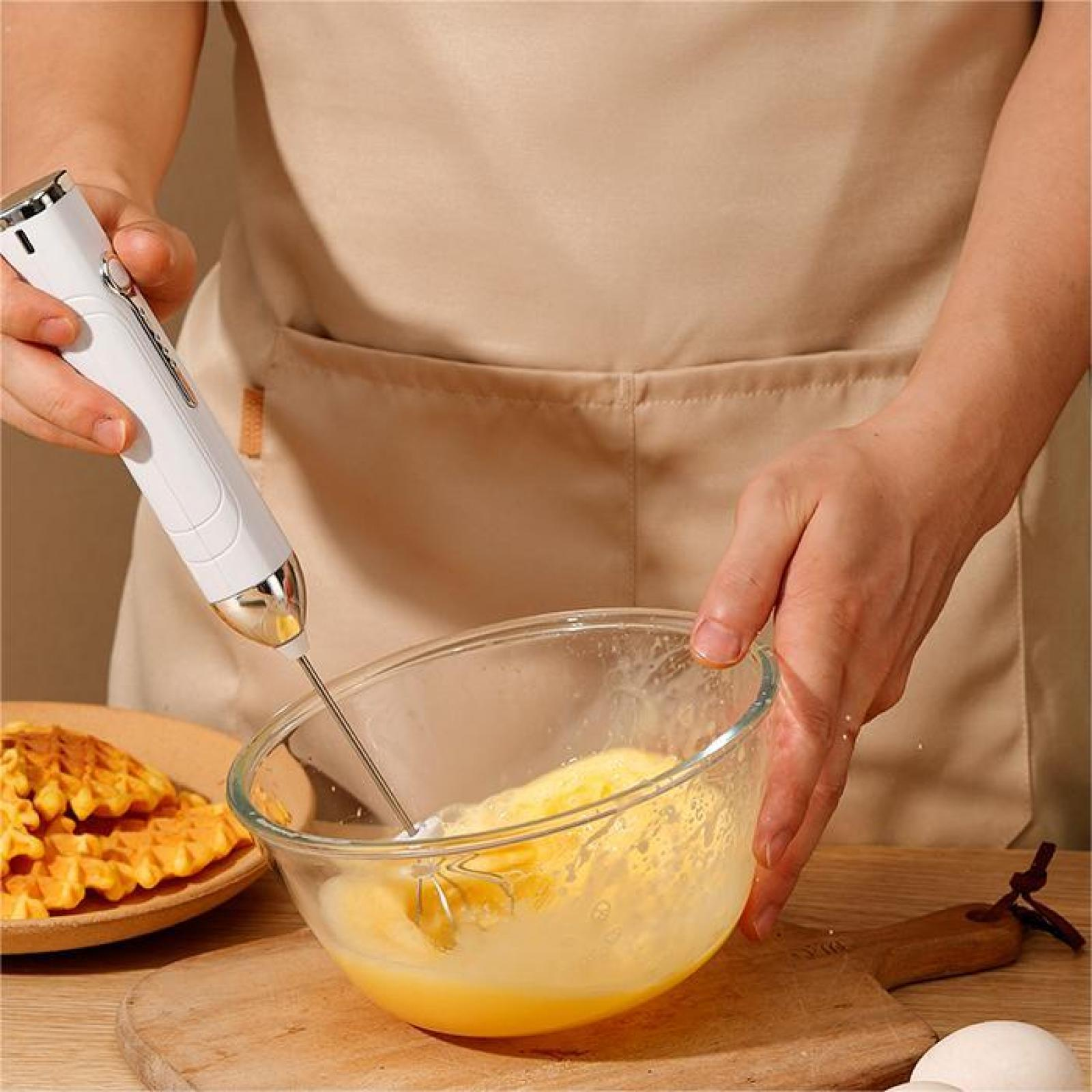 Eine Person verrührt Eier in einer Glasschüssel mit einem Handmixer. Waffeln sind im Hintergrund auf einem Teller.