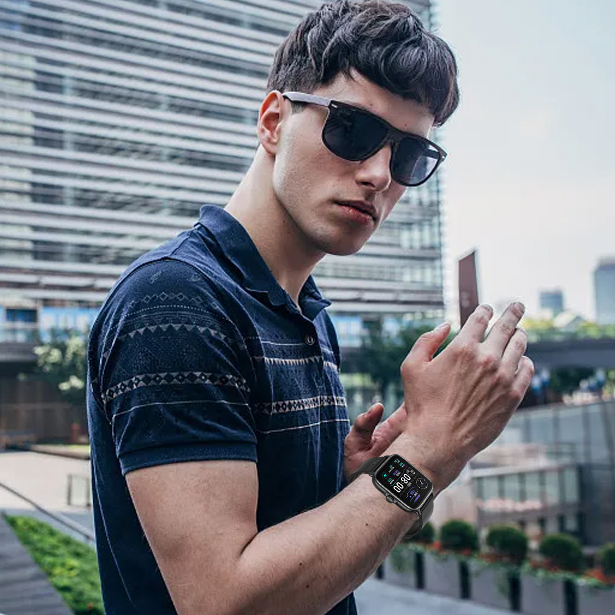 Ein Mann mit Sonnenbrille und Smartwatch posiert draußen. Gebäude und Grünflächen sind im Hintergrund zu sehen.