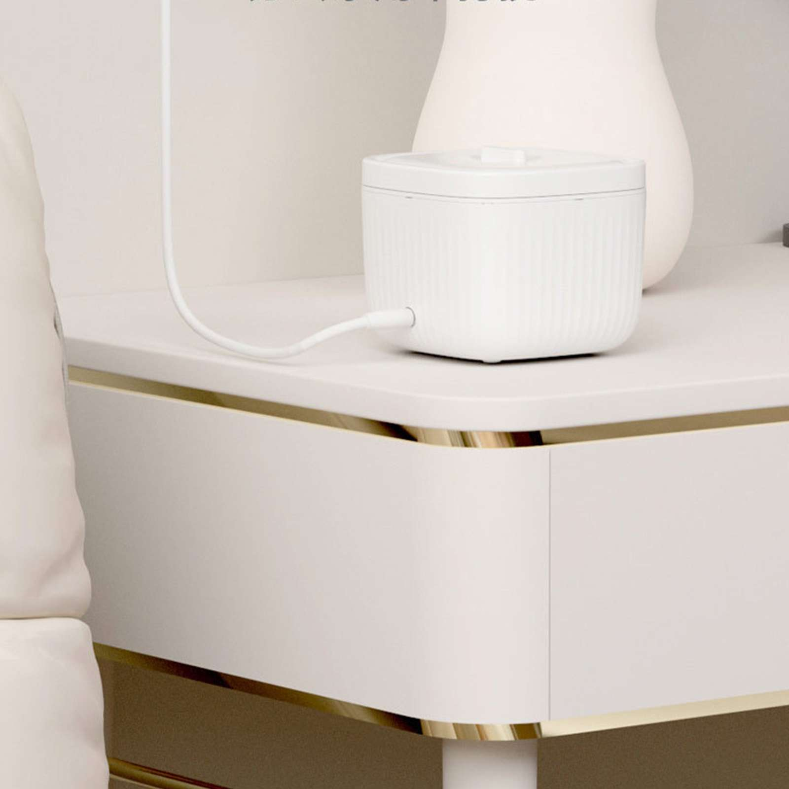 Weißes Gerät mit Kabel auf einem weißen Tisch mit goldenen Zierleisten. Im Hintergrund eine weiße Vase.