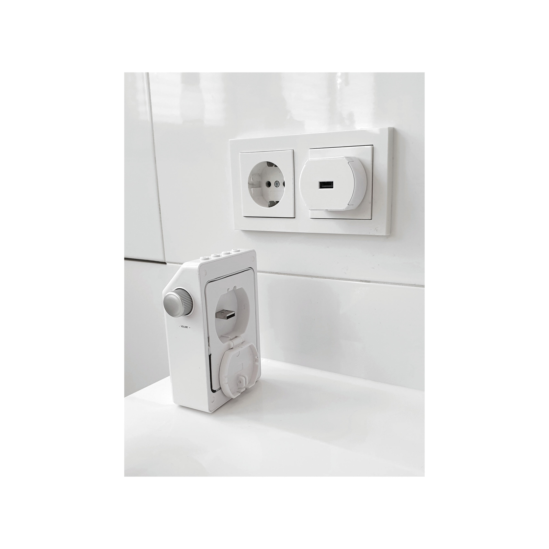 Weiße Steckdose mit USB-Ladegerät und einem Lautstärkeregelungsgerät daneben, alles auf einer weißen Fliesenoberfläche.