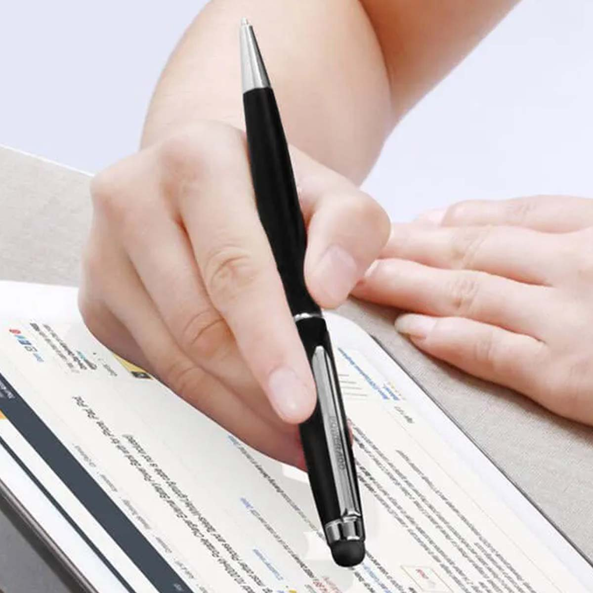 Una mano sostiene un bolígrafo negro sobre una tableta. El bolígrafo tiene punta y clip plateados. La tableta muestra texto y el fondo está borroso.