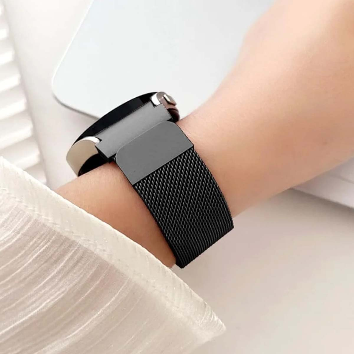 Il polso di una persona con uno smartwatch nero. Il cinturino è in rete tessuta. La persona indossa una camicia bianca.