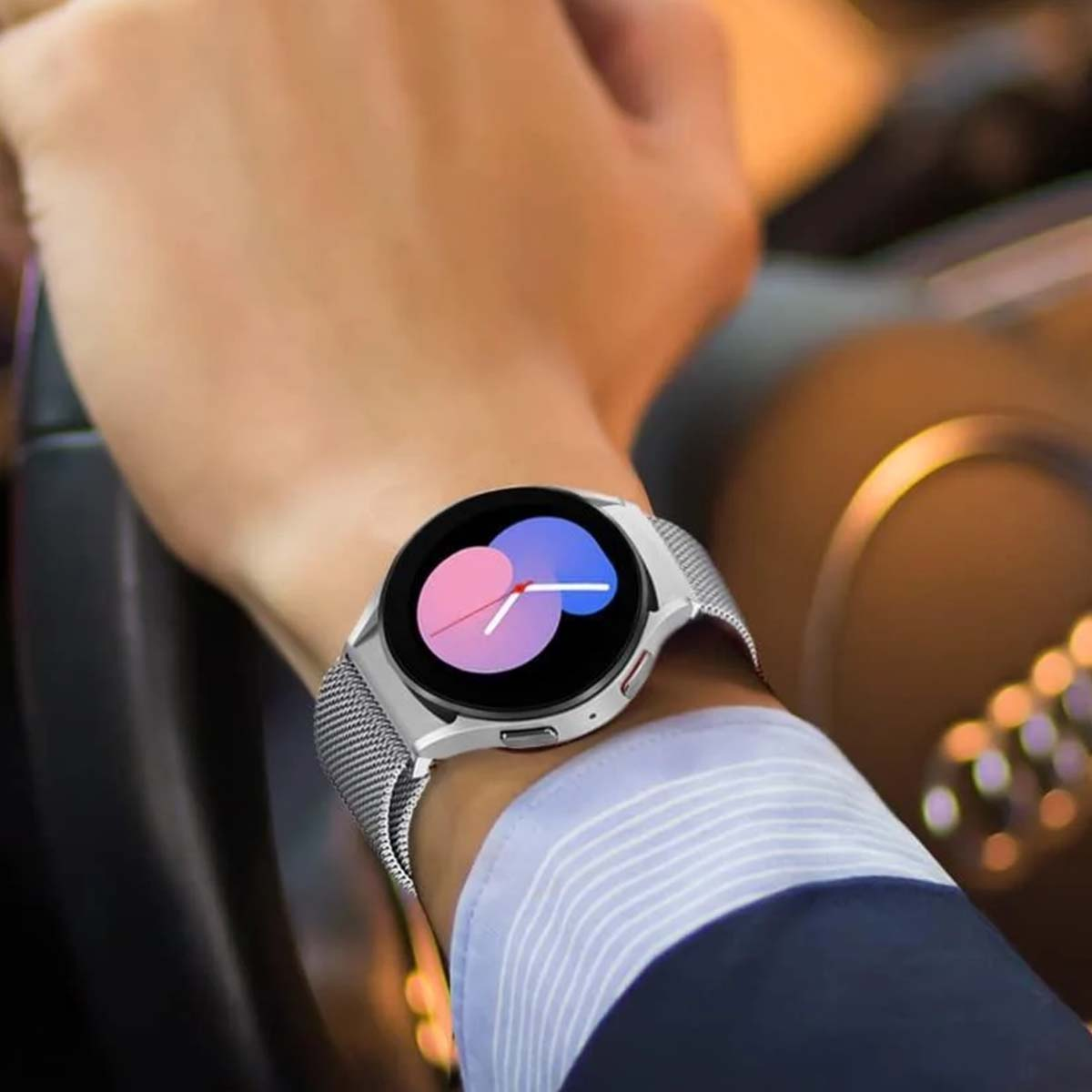 Una persona indossa uno smartwatch argentato con schermo rosa e blu. L'orologio è sul polso sinistro. Sta guidando un'auto.
