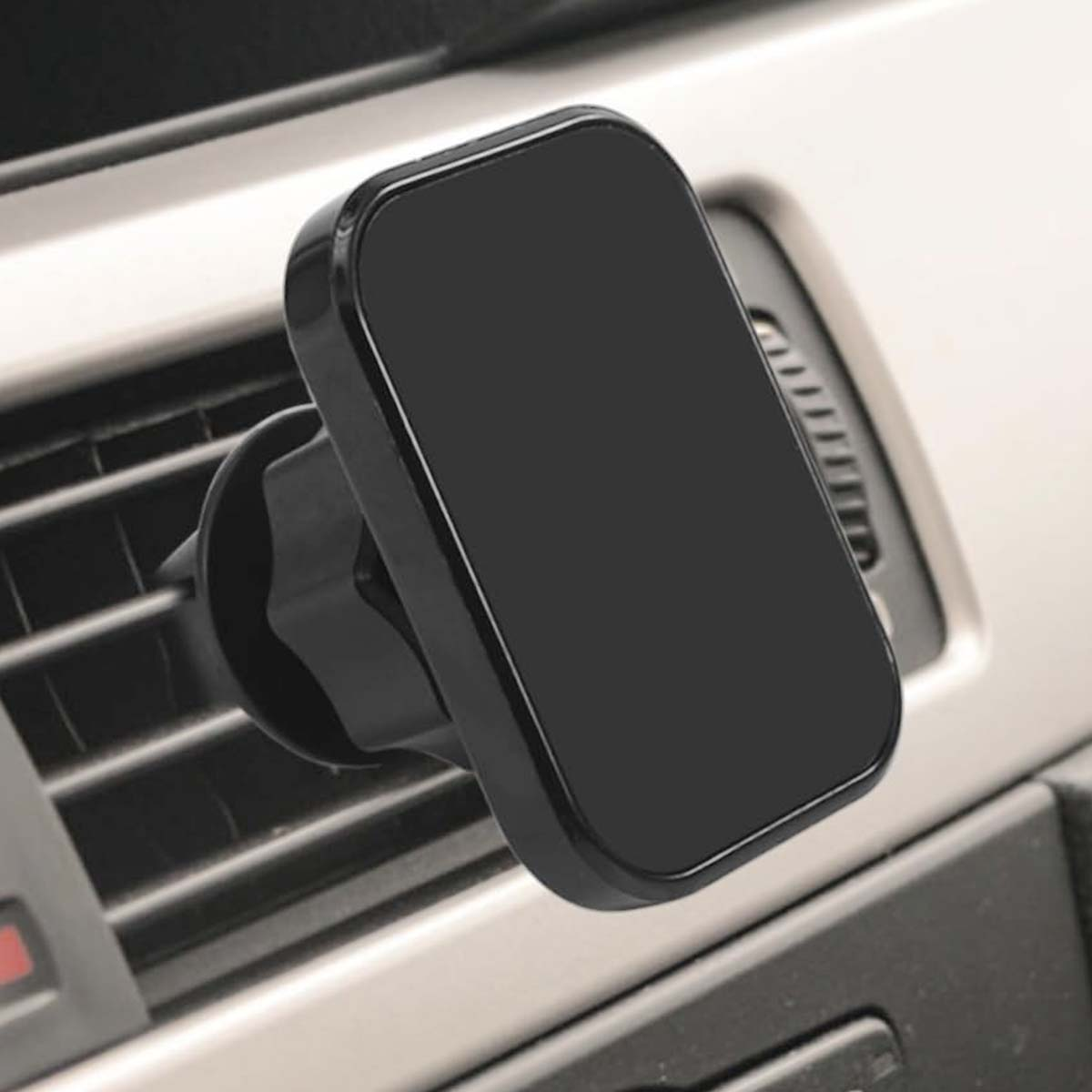 Un soporte negro para teléfono está montado en una rejilla de ventilación del coche. El soporte tiene forma rectangular con esquinas redondeadas. El interior del coche es visible.