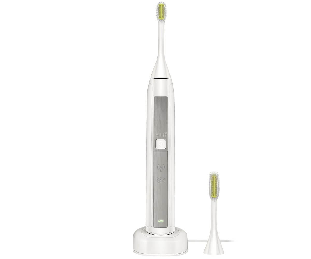 Une brosse à dents électrique blanche avec une base de chargement et une tête de brosse supplémentaire. La brosse a un panneau argenté et une tête verte.