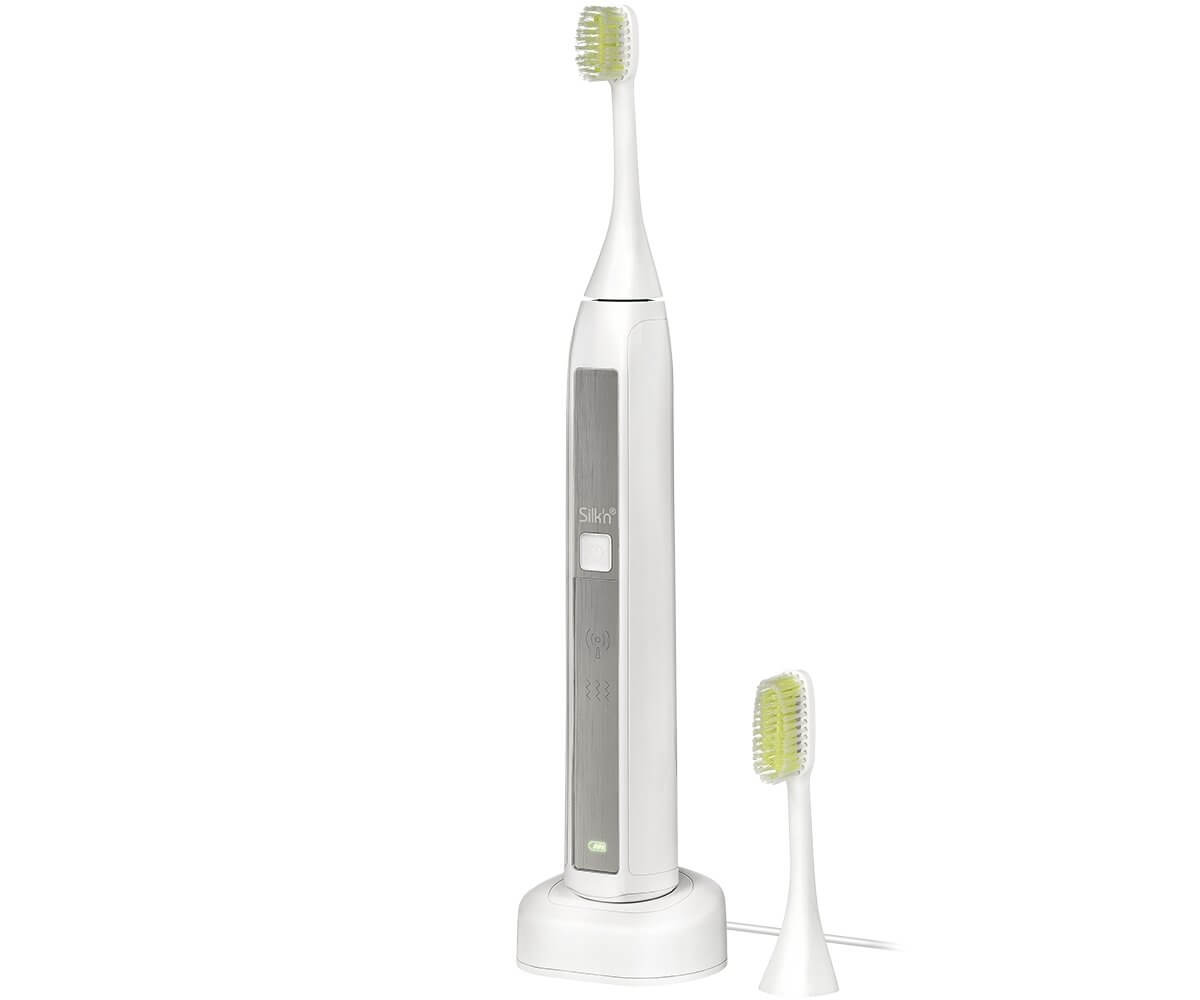 Brosse à dents électrique blanche avec des poils verts, et une tête de rechange. Elle est sur un socle de chargement, sur une surface blanche.