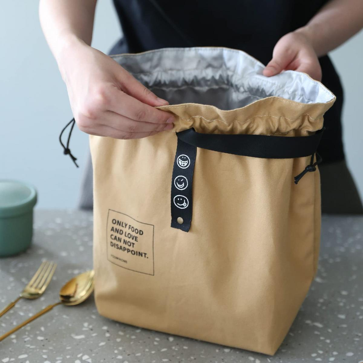 Eine Person öffnet eine beige Lunchtasche mit schwarzem Riemen und Smiley-Gesichtern. Text: Nur Essen und Liebe können nicht enttäuschen.