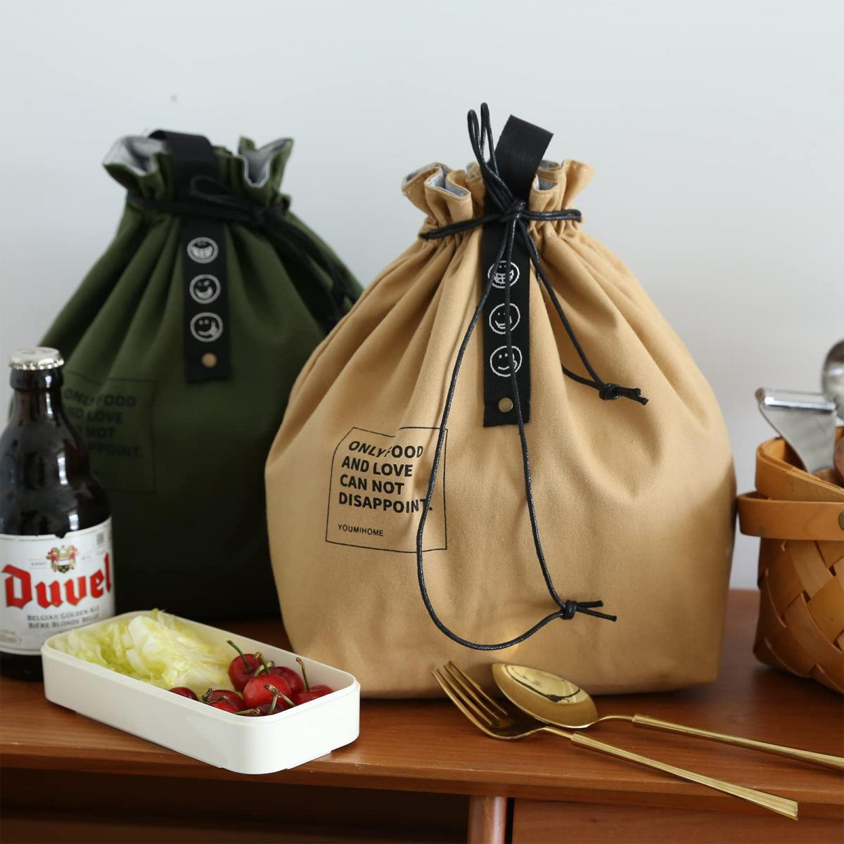 Zwei Lunchbags, einer grün, einer beige, mit einer Bierflasche, einem Lebensmittelbehälter und Besteck auf Holz.