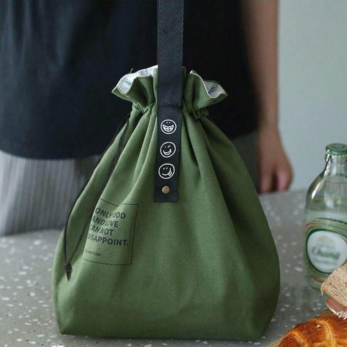 Grüne Lunch-Tasche mit Smiley-Gesichtern und einem Zitat. Im Hintergrund eine Person, auf einem Tisch eine Wasserflasche und Brot.