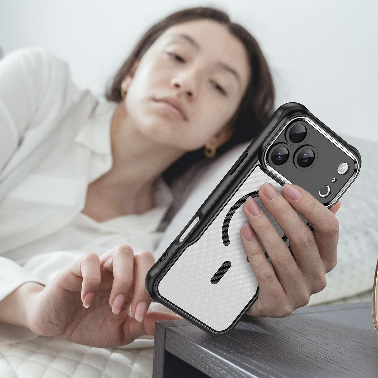 Eine Frau im Bett hält ein Telefon mit einer schwarz-weißen Hülle. Das Telefon hat drei Kameras. Sie trägt ein weißes Hemd.