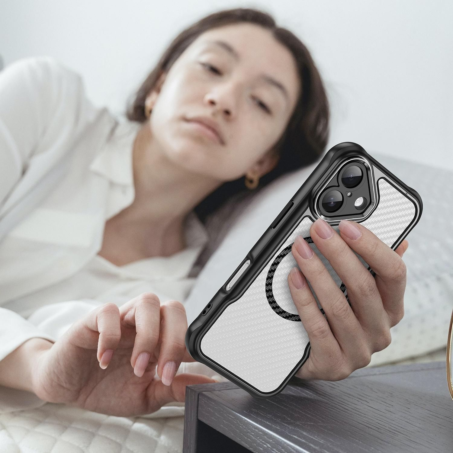 Eine Frau im Bett hält ein Telefon in einer Hülle. Das Telefon befindet sich in der Nähe des Nachttisches. Sie trägt ein weißes Hemd und hat goldene Ohrringe.