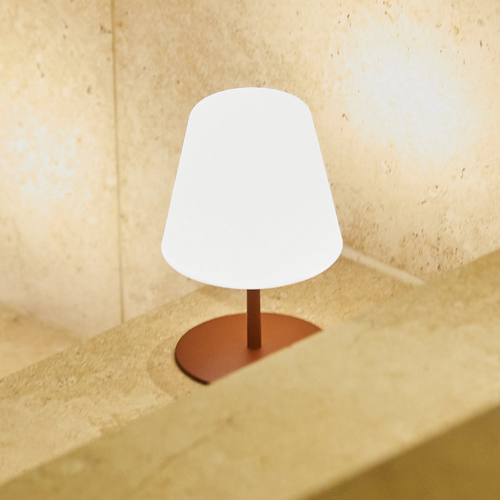 Eine kleine weiße Lampe mit rotem Fuß steht auf einer Steinoberfläche. Der Hintergrund ist eine beige Steinmauer.