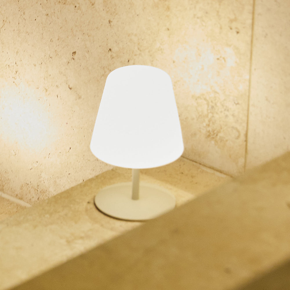 Eine kleine weiße Lampe steht auf einer Steinoberfläche vor einer strukturierten Wand. Die Lampe ist beleuchtet, im Vordergrund ist ein Holzbalken.