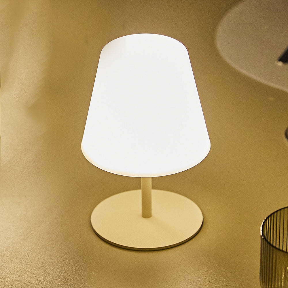 Eine kleine, beleuchtete weiße Lampe mit konischem Schirm steht auf einem Tisch. Die Lampe hat einen kreisförmigen Fuß und einen dünnen Stiel.