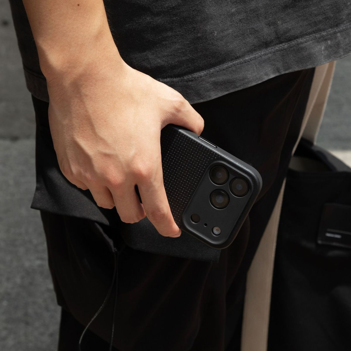 Die Hand einer Person hält ein schwarzes Telefon mit einer strukturierten Hülle. Sie trägt schwarze Kleidung. Im Hintergrund ist eine Tasche zu sehen.