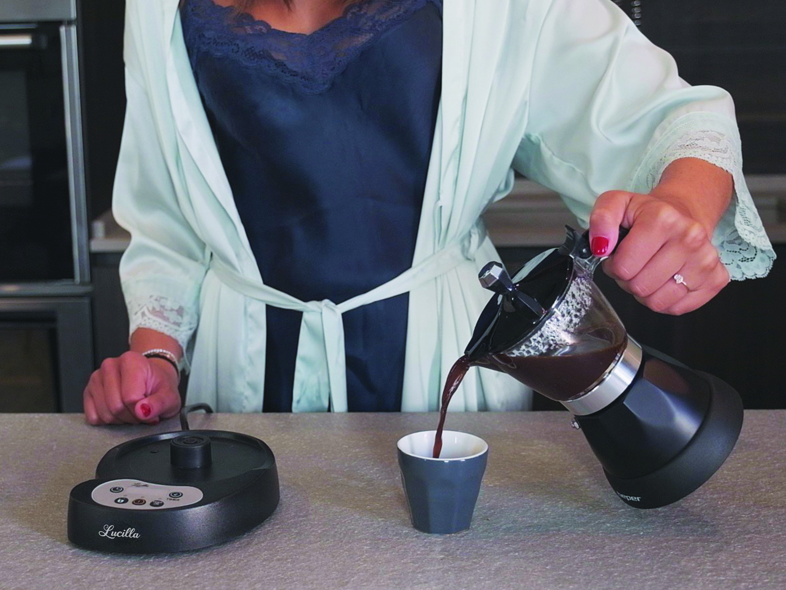 Una persona versa caffè da una caffettiera moka nera in una tazza grigia. Un scaldavivande nero a sinistra.