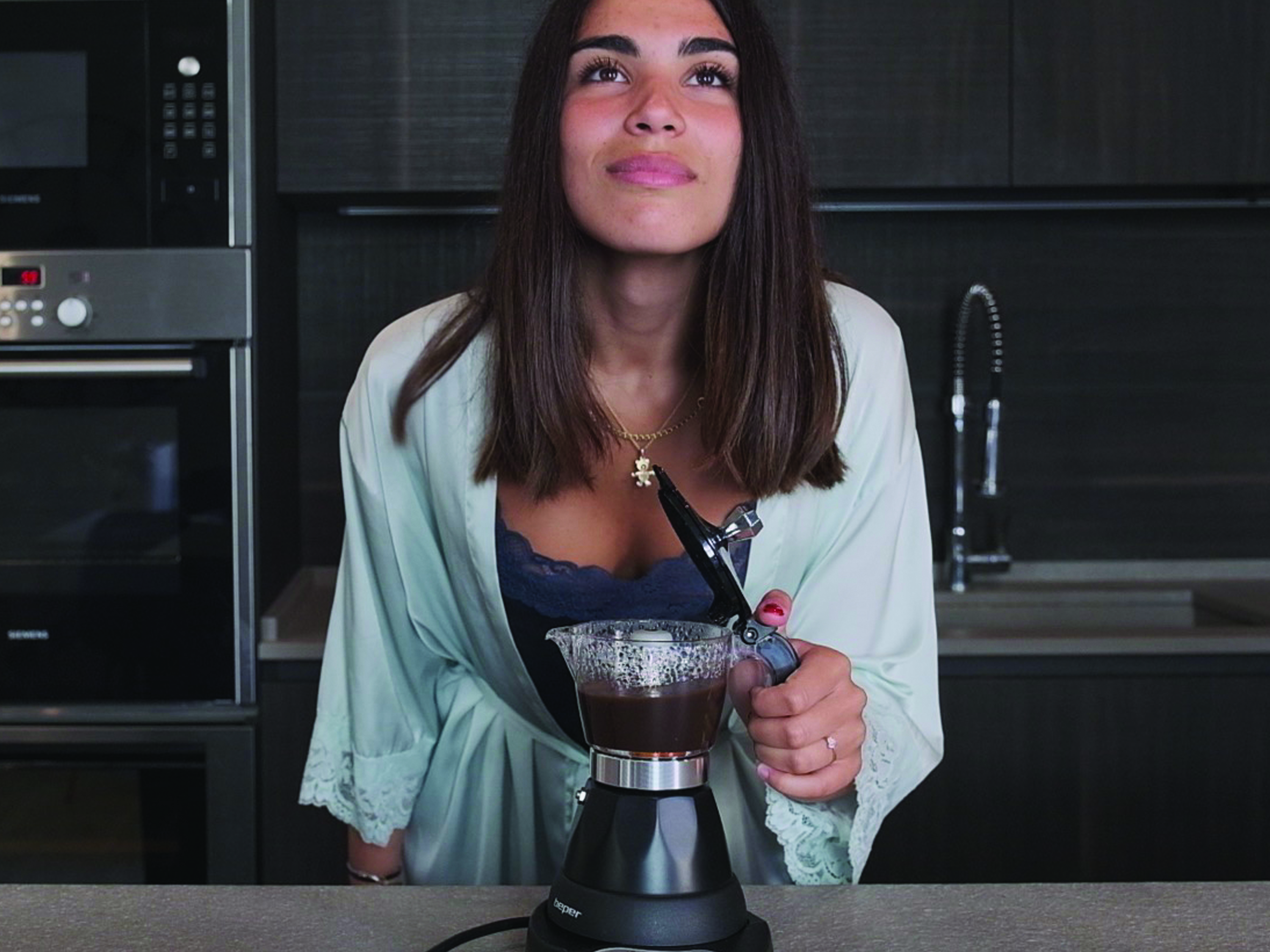 Donna in accappatoio che prepara il caffè con una moka. Guarda in alto. Elettrodomestici da cucina sullo sfondo.