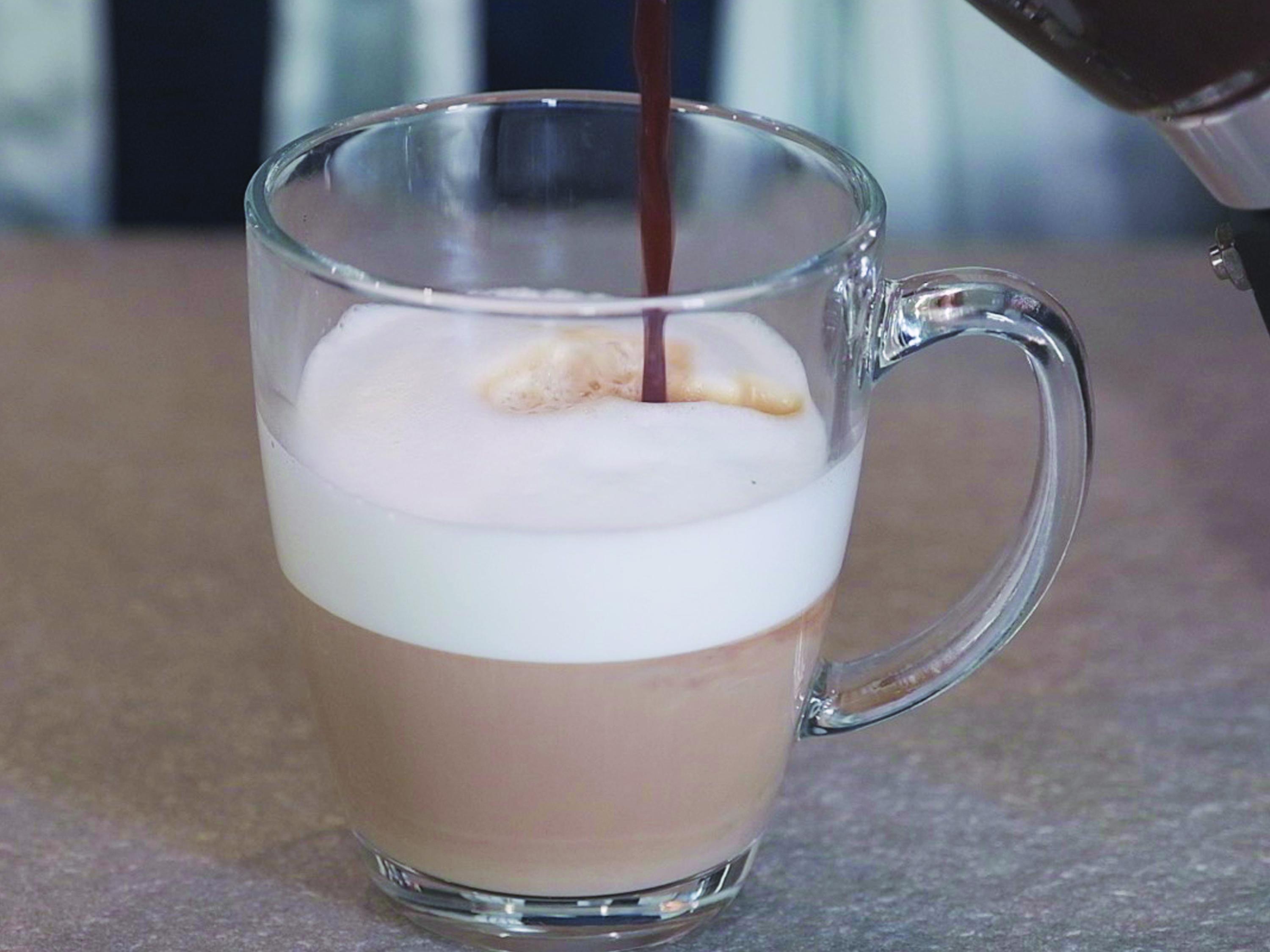 Caffè versato in una tazza di vetro trasparente con strati di schiuma di latte e caffè. La tazza è su un tavolo.