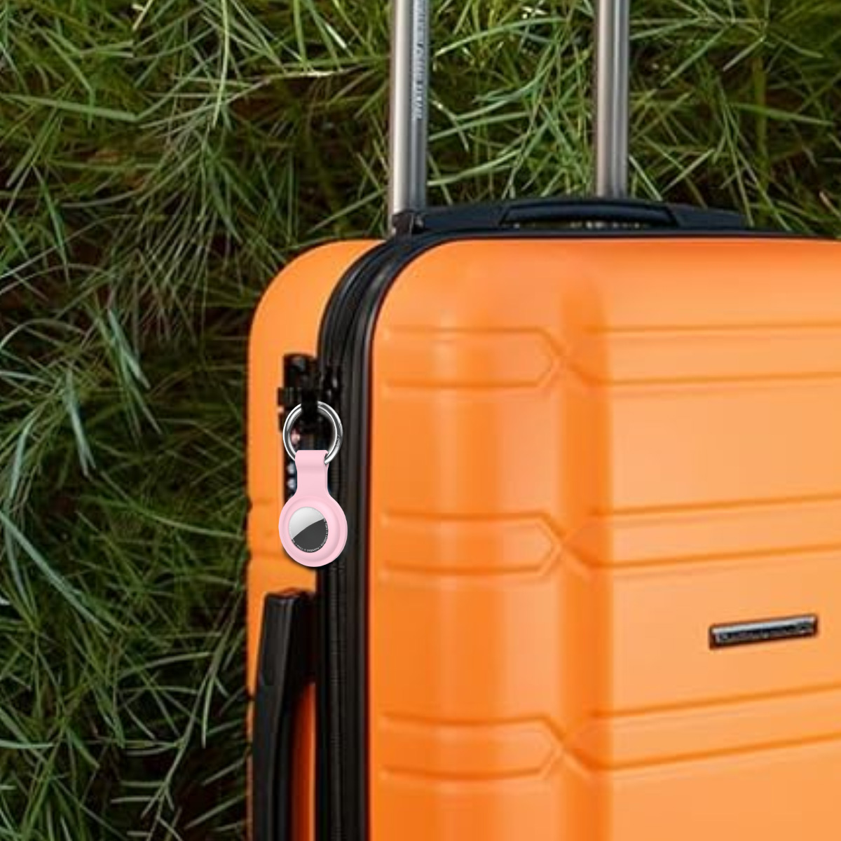 Ein orangefarbener Koffer mit einem rosa Tracker. Im Hintergrund ist grünes Gras zu sehen. Der Koffer hat einen schwarzen Griff und silberne Stangen.