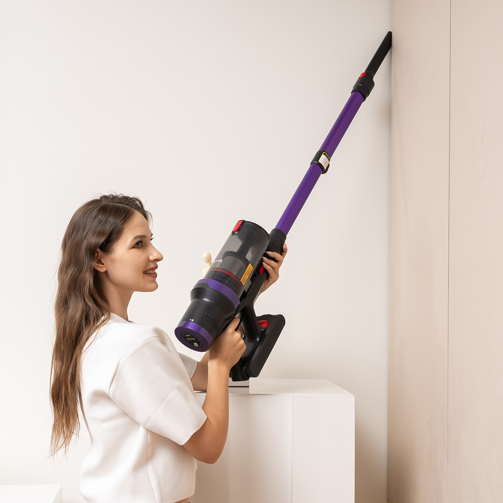 Kobieta w białej bluzce trzyma fioletowo-czarny odkurzacz, czyszcząc biały róg ściany.