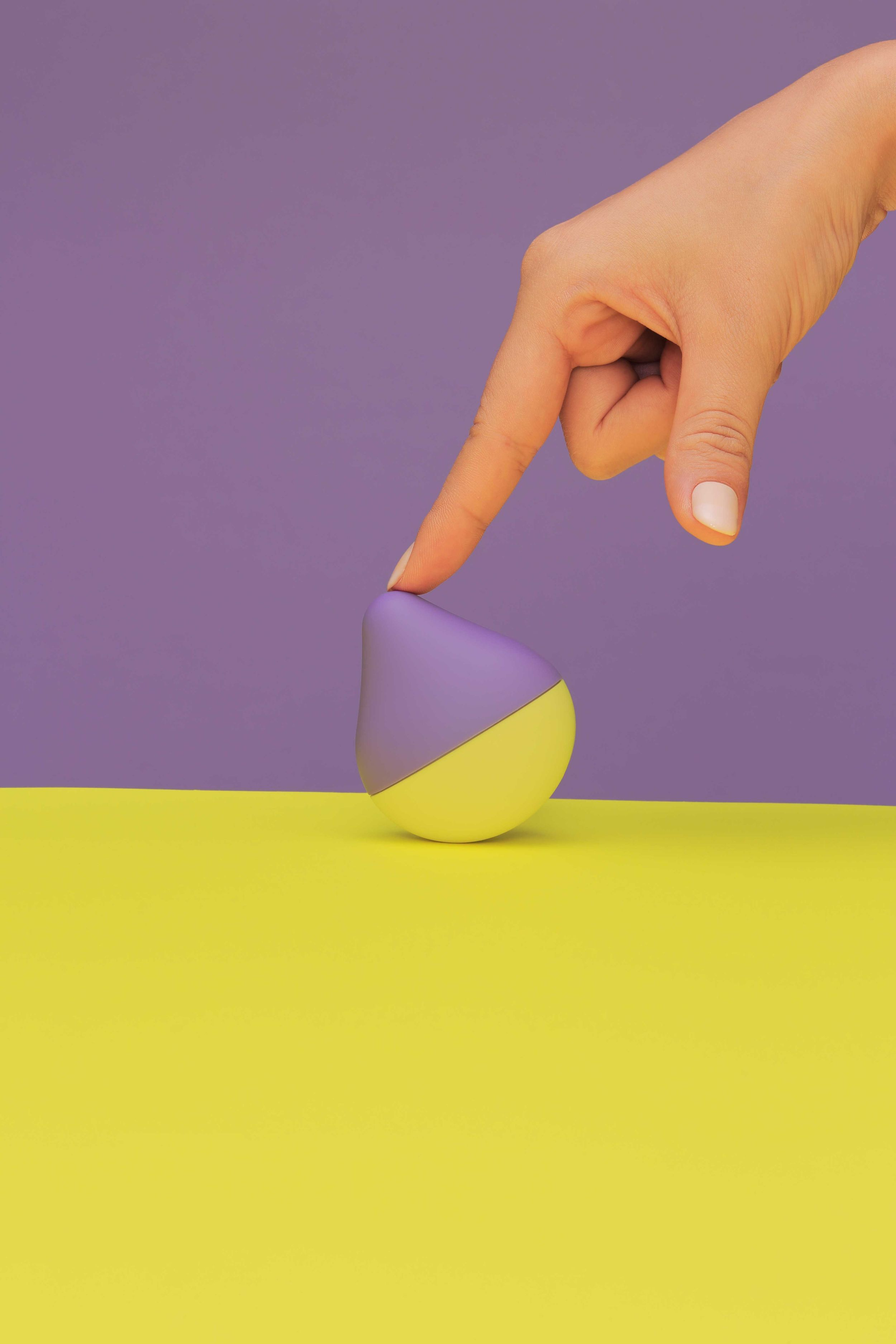 Dłoń dotykająca fioletowego i żółtego przedmiotu. Fioletowe tło i żółta powierzchnia. Palec wskazujący w dół.