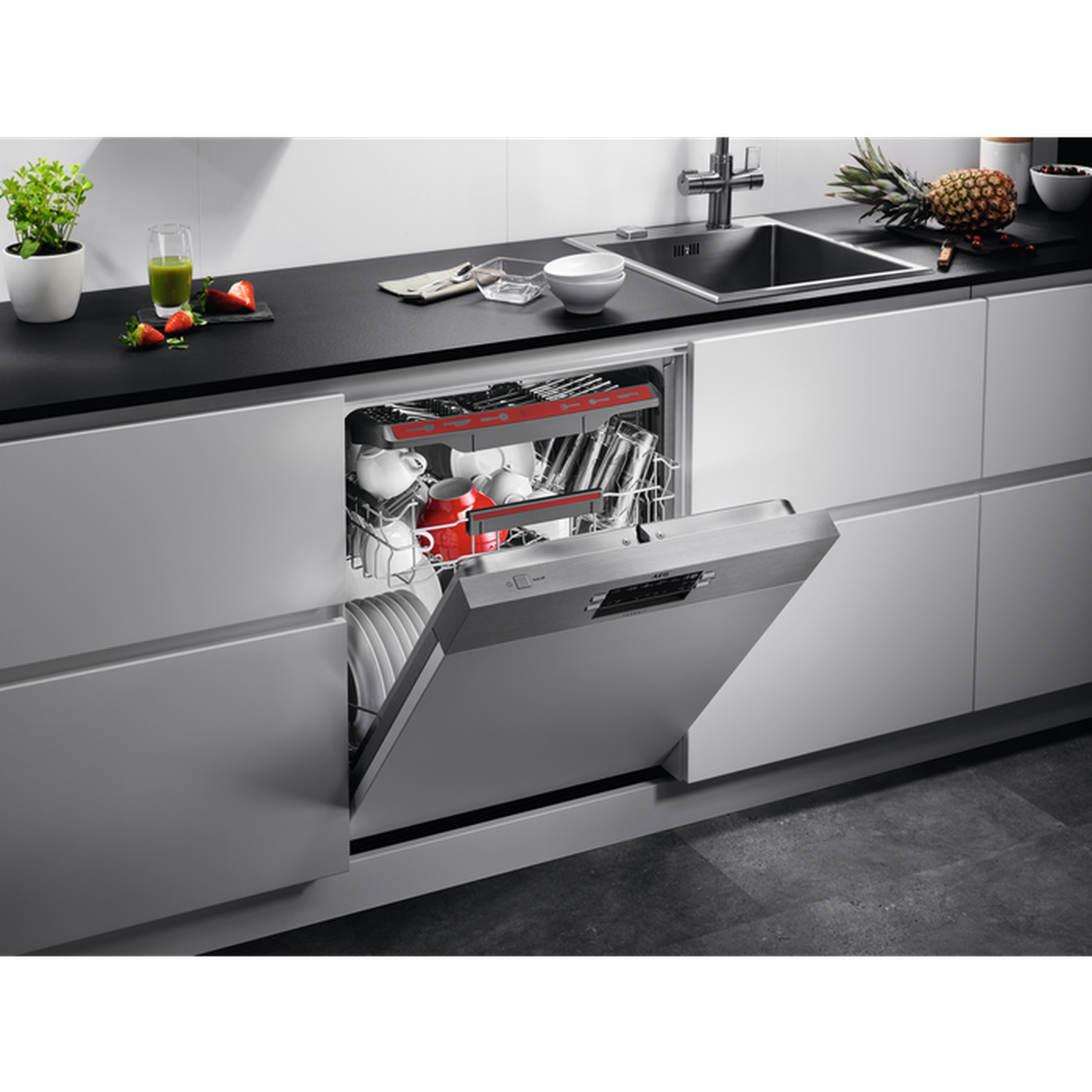 An open dishwasher filled with dishes. A kitchen counter has a sink, fruit, and plants.